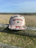 Camouflage cap with 'Northwoods Outfitters' logo on a wooden post with a field and blue sky in the background