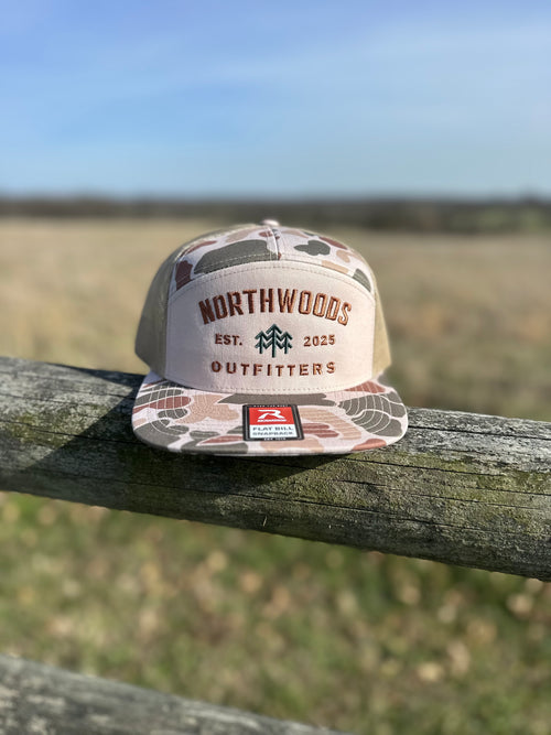 Camouflage cap with 'Northwoods Outfitters' logo on a wooden post with a field and blue sky in the background